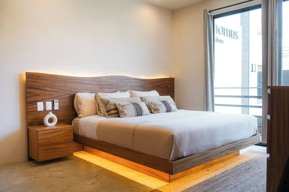 A modern bedroom with a wooden headboard, a bedside table with a lamp, and a large window.