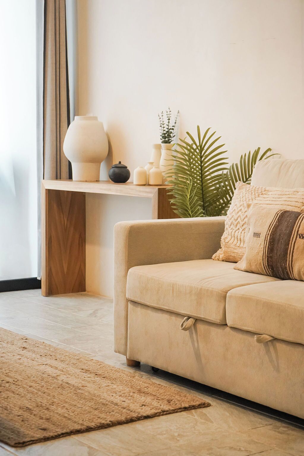 A beige sofa with a textured throw pillow, a small wooden side table with decorative items, a large green plant, and a beige rug in a modern living room.