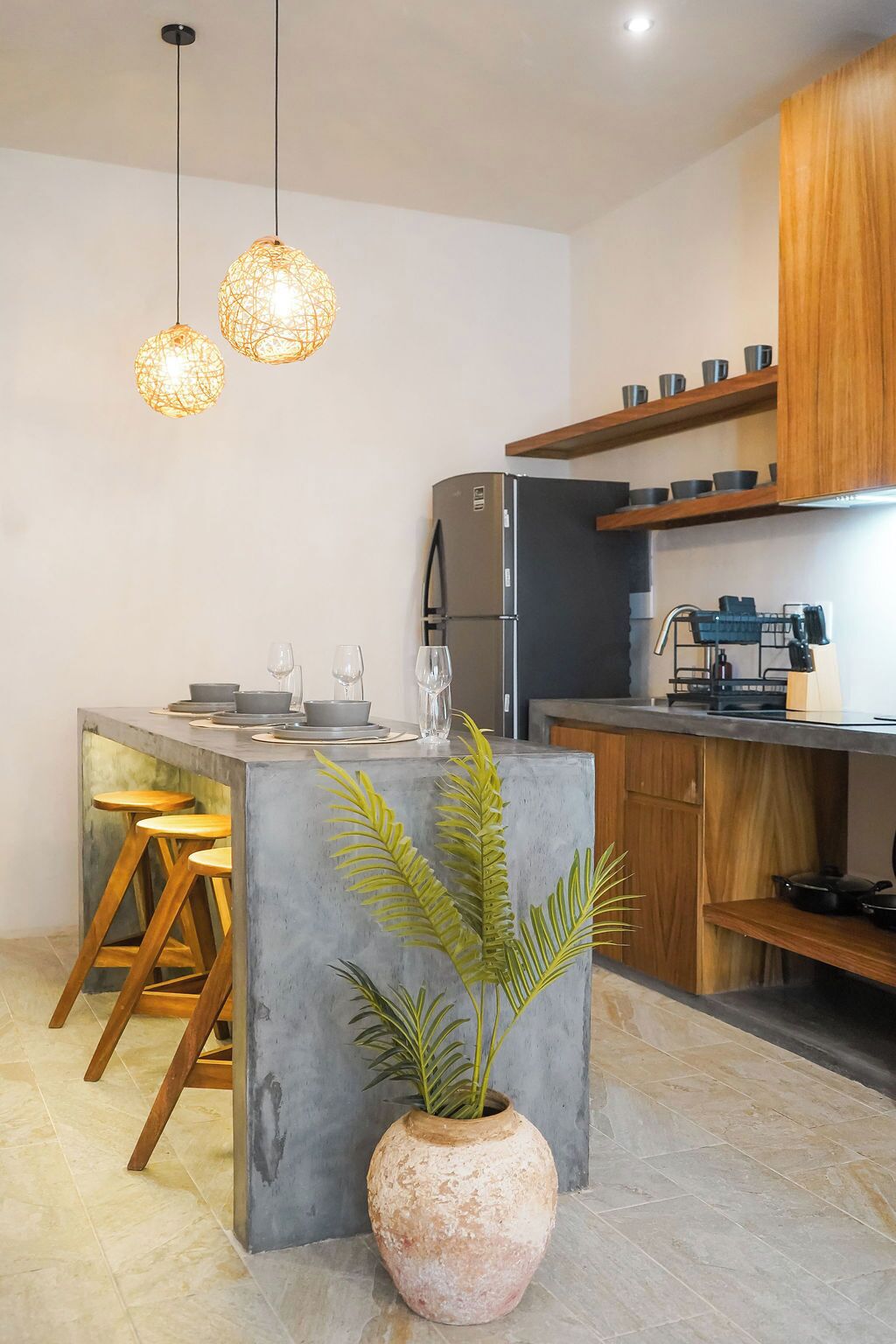 A modern kitchen with a central island, wooden stools, a potted plant, hanging pendant lights, and built-in wooden shelves.