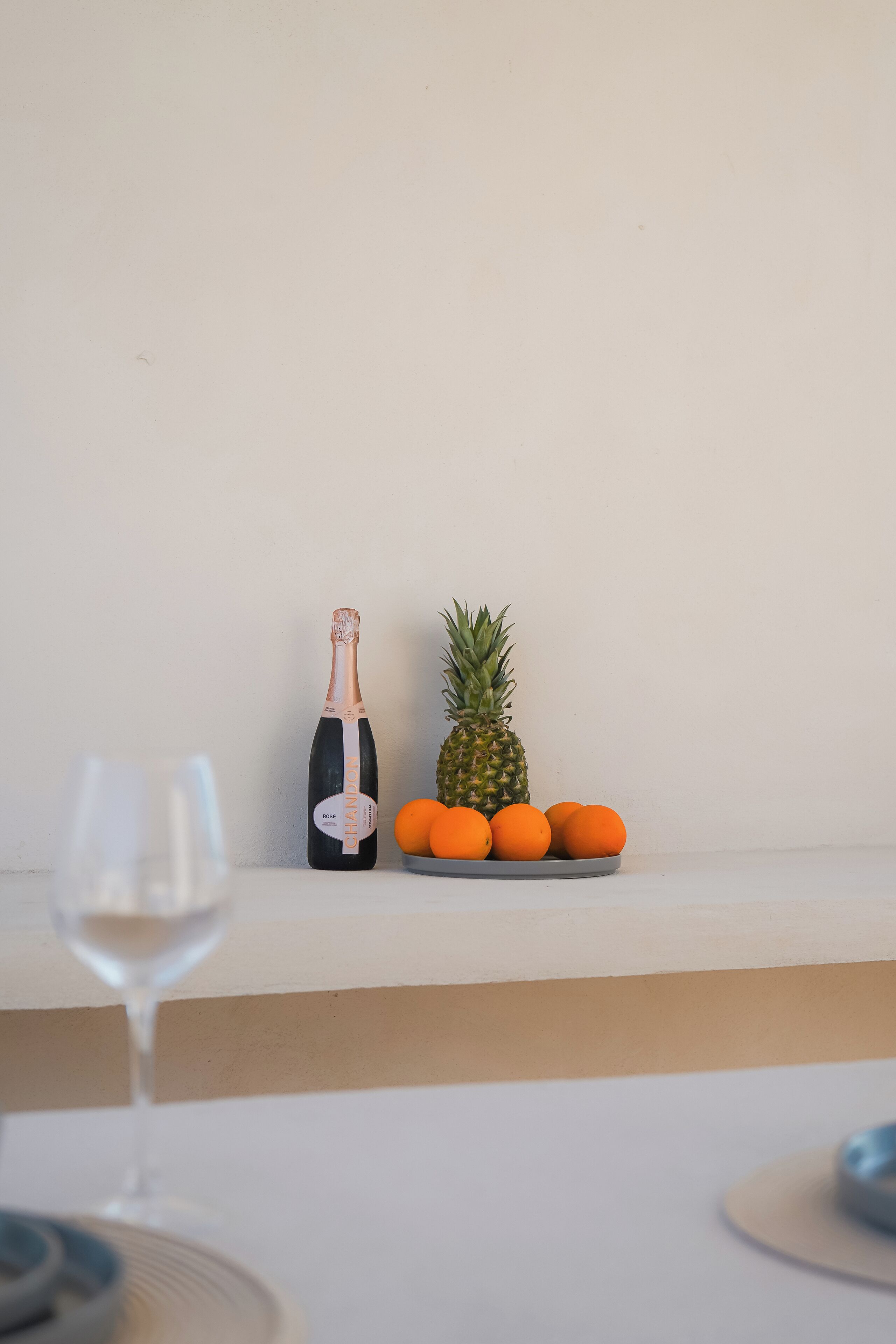 Una botella de vino Chandon, una piña y naranjas sobre una mesa frente a una pared lisa.