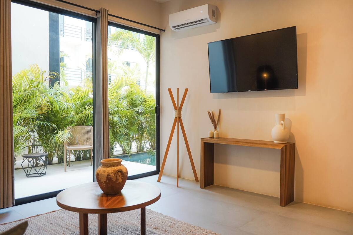 A modern living room with a large window, a flat-screen TV mounted on the wall, a wooden console table, and a rustic vase on a round table.