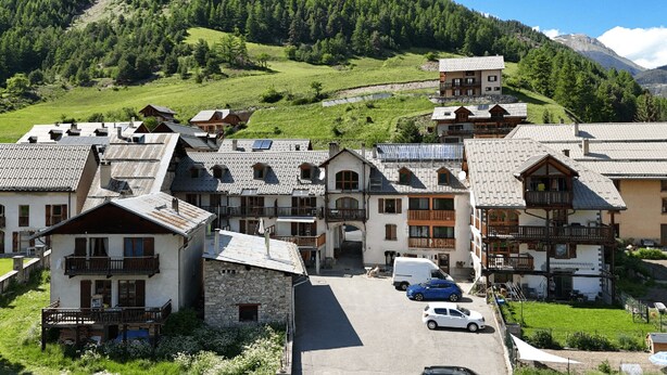 A cluster of traditional chalet-style buildings with stone facades and wooden balconies, set against a backdrop of lush green hills and a clear sky. - Gites Simond welcomes you to Arvieux in the heart of the Queyras Regional Park, (Arvieux)