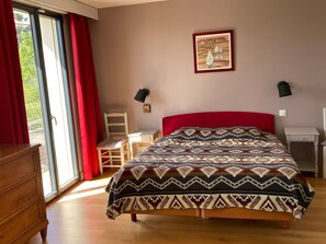A bedroom with a bed, a chair, a small table, a dresser, a window with curtains, a wall-mounted lamp, and a framed picture.