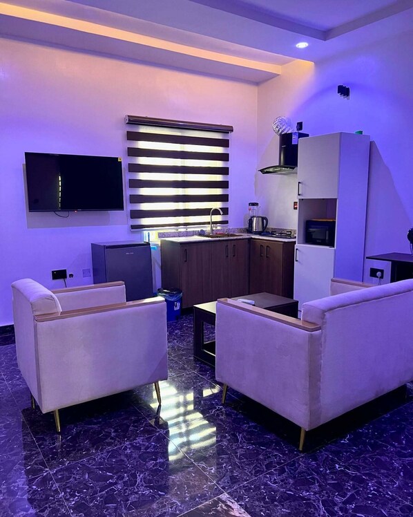A modern living room with a flat-screen TV, two sofas, a coffee table, and a kitchen area with cabinets and a stove. - Triple Homes Apartment (Abuja)