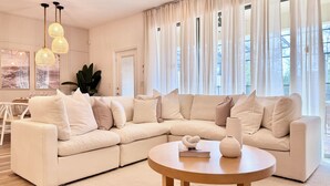 A modern living room with a beige sectional sofa, a round wooden coffee table, and a large window with sheer curtains.