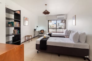 A modern hotel room with two beds, a desk, a bookshelf, and a view of the city through the window. - Comfort and Tranquillity in Humait M404 (Rio de Janeiro)