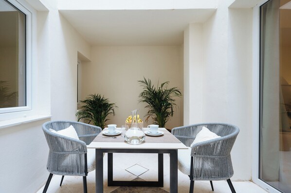 A small, well-lit dining area with a table set for two, surrounded by wicker chairs, with potted plants and a clear view of the outside. - Luxurious Apartment with Patio and Pool (Seville)