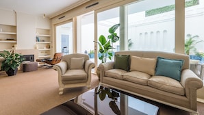 Ein geräumiges Wohnzimmer mit einem beigen Sofa, einem Holzsesseln und einem Couchtisch. Große Fenster bieten einen Blick ins Freie.