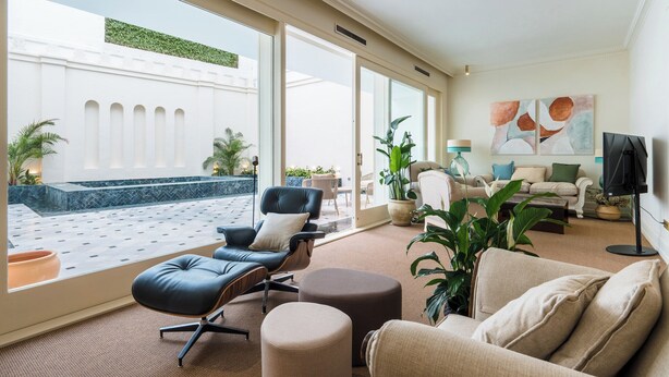 Ein geräumiges Wohnzimmer mit großem Fenster, einem schwarzen Ledersessel, einem beige farbenen Sofa und einem Flachbildfernseher.