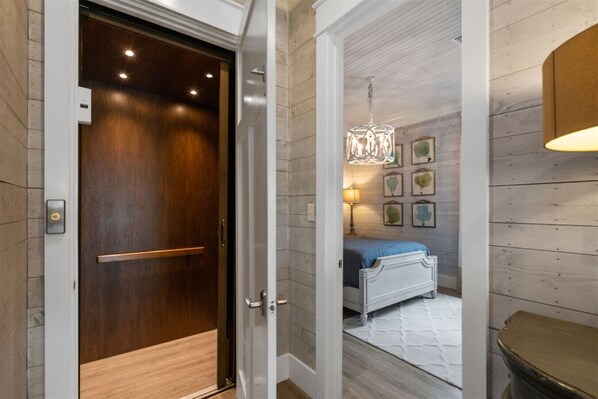 A hotel room with a wooden door, a bed with a blue bedspread, a white headboard, a nightstand with a lamp, and a wall with framed pictures. - Cottages of Romar 12 - Beach Please - Luxury Home (Orange Beach)
