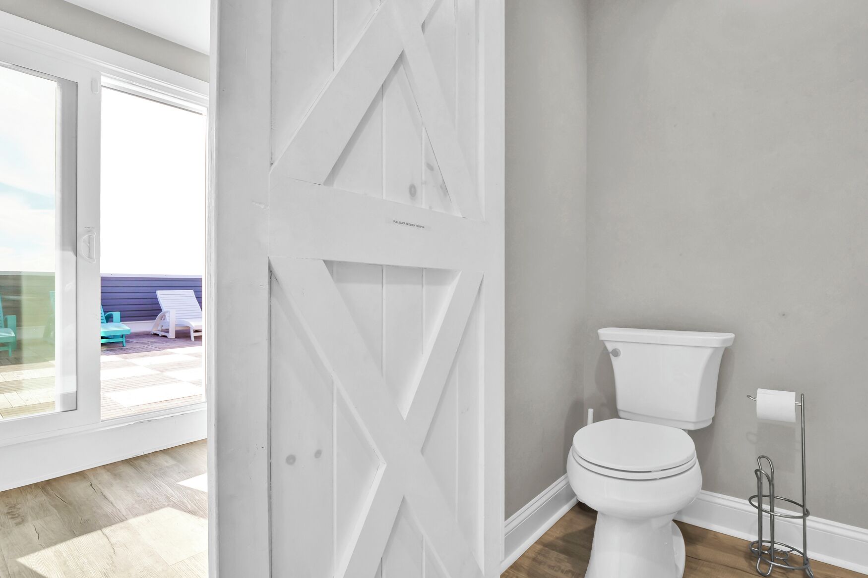 A bathroom with a white toilet, a white door with a geometric pattern, and a view of a deck with outdoor furniture.