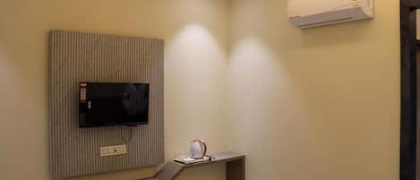 A hotel room with a flat-screen TV mounted on a wall, a desk with a lamp, and an air conditioning unit.