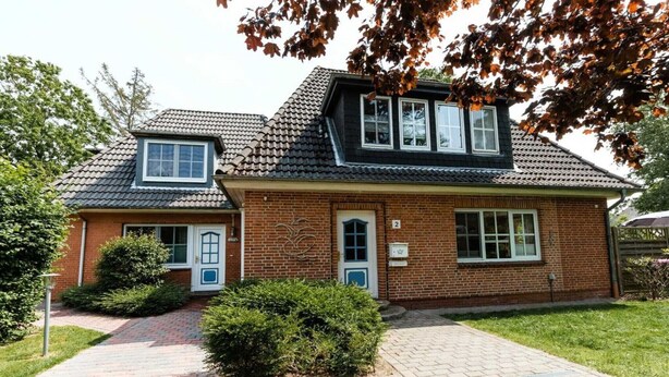 Une maison à deux étages, construite en briques, avec un toit en tuiles, un petit garage attenant et une allée pavée.