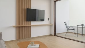 A modern living room with a flat-screen TV mounted on a wooden panel, a small table with a book and a mug, and a view of a balcony with a chair and table.