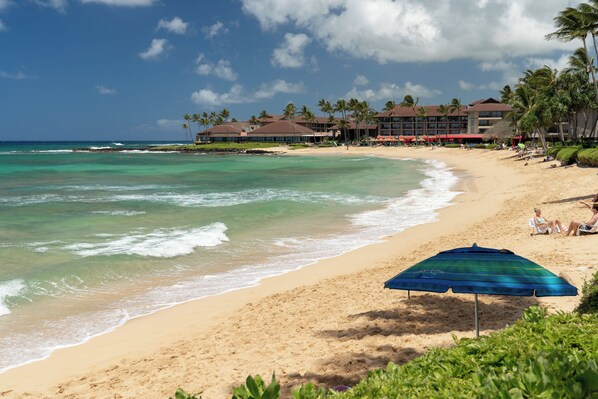 A beach with clear turquoise waters, a sandy shore, and a resort building with multiple balconies and palm trees.