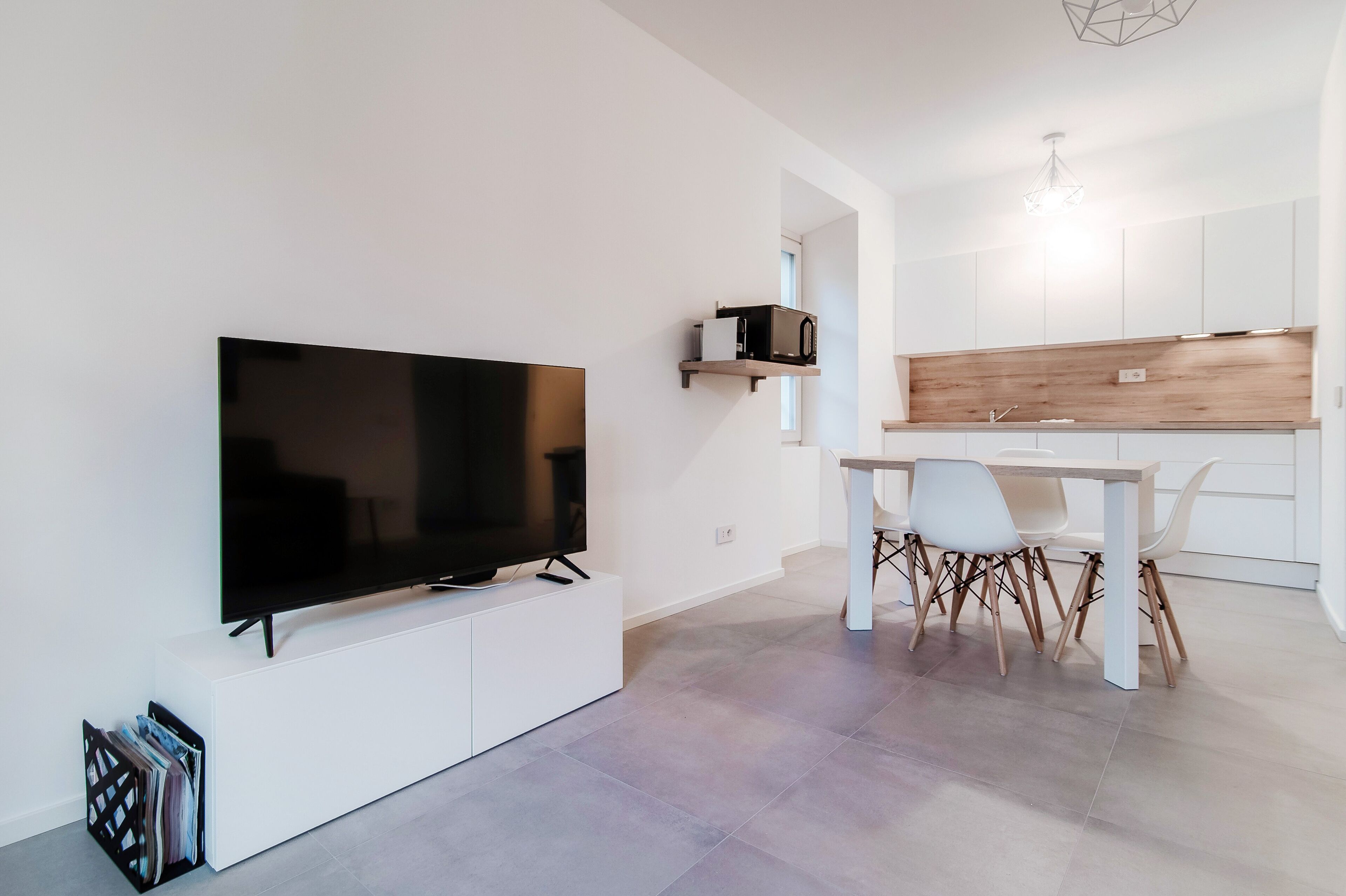 Un espace de vie moderne doté d’un téléviseur à écran plat fixé sur un meuble blanc, d’un coin repas avec une table et des chaises blanches, et d’une cuisine avec des meubles blancs et un pare-feu en bois.