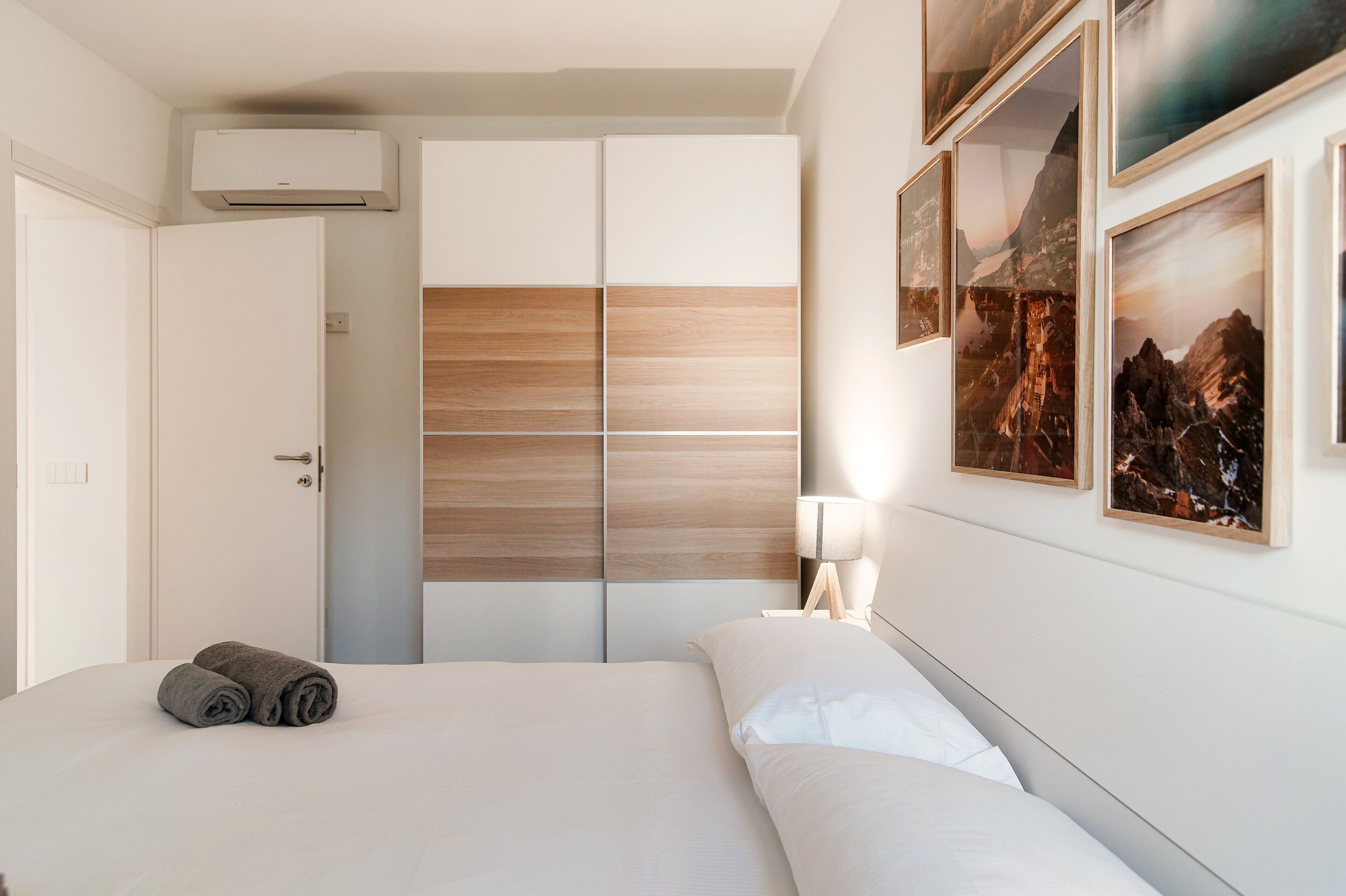 A hotel room with a bed, a bedside table with a lamp, a wardrobe with a wooden panel design, and framed pictures on the wall.