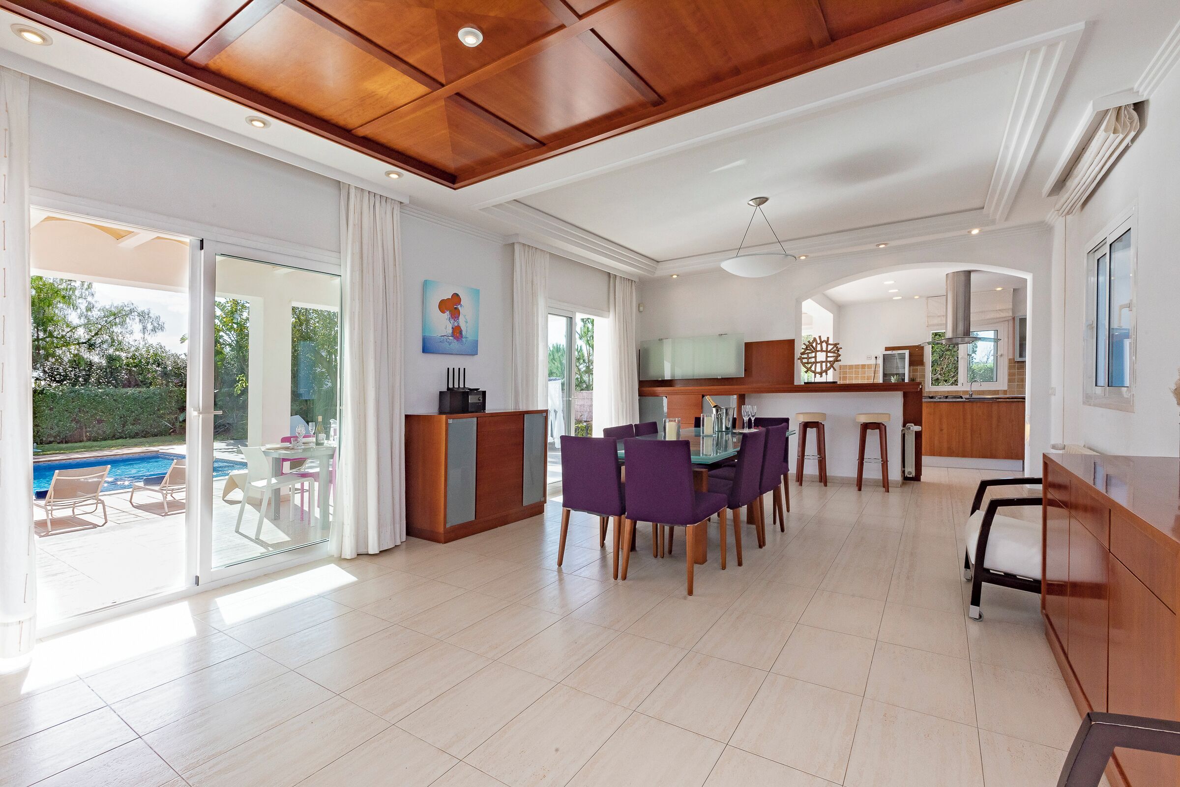 Amplia sala de estar con una mesa de comedor, sillas moradas, una cocina y un ventanal corredizo que da a una piscina.