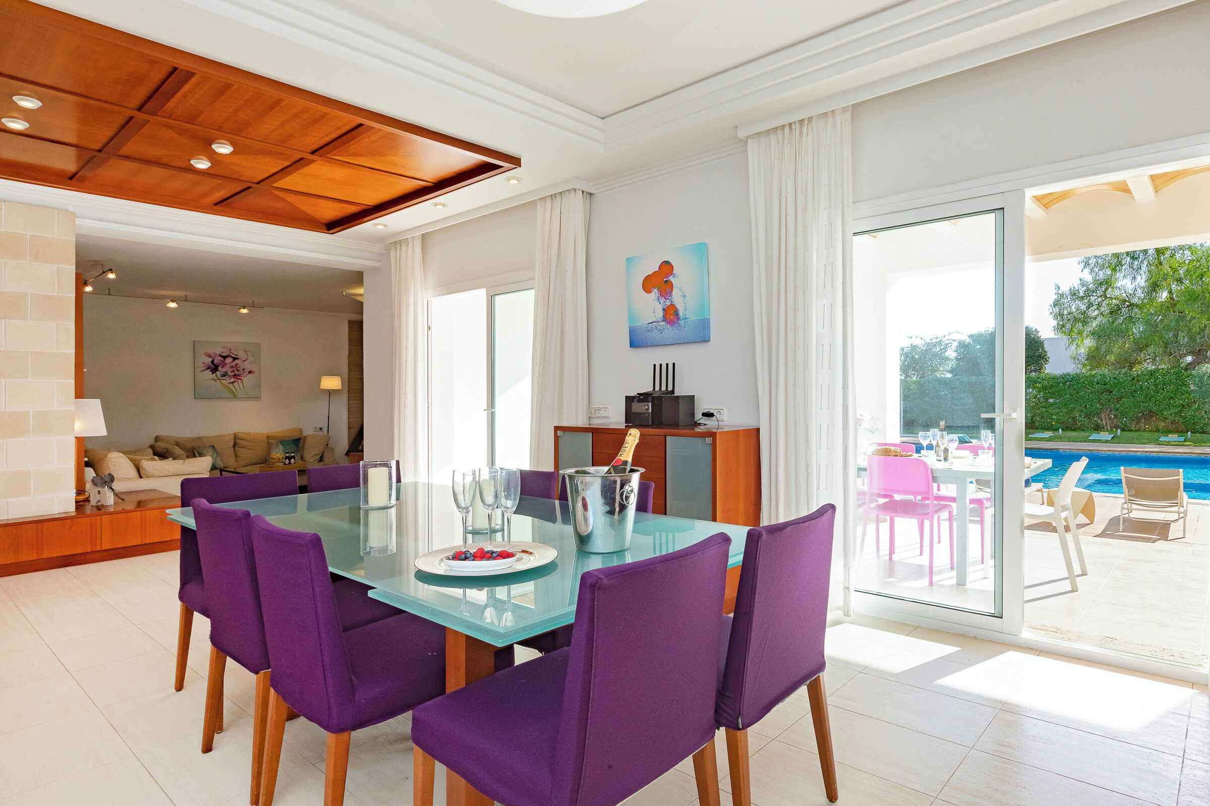 A modern dining area with a glass table and purple chairs, a kitchen in the background, and a view of a swimming pool.