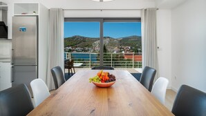 Un espace de repas moderne avec une table en bois, des chaises et un saladier rempli de fruits. Une grande fenêtre offre une vue sur une ville côtière et des collines.