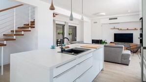 Une cuisine moderne dotée d’un îlot central, un coin repas avec un canapé gris, un téléviseur et un escalier avec des marches en bois.