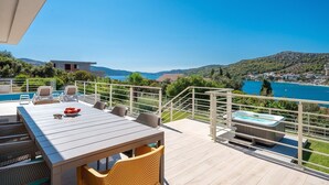 Une terrasse dotée d’une piscine, de chaises longues, d’une table avec un saladier rempli de fruits et d’un jacuzzi, offrant une vue imprenable sur un lac pittoresque et des collines.