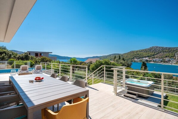 A terrace with a pool, lounge chairs, a table with a bowl of fruit, and a hot tub, overlooking a scenic lake and hills.
