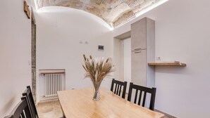 A dining area with a wooden table, chairs, a vase with dried pampas grass, a radiator, and a brick ceiling.