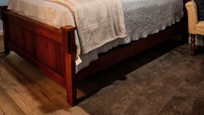 A wooden bed with a headboard, a bedside lamp, and a chair in a room with wooden flooring and a sloped ceiling.