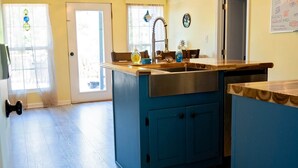 A kitchen with a blue island, wooden ceiling beams, a ceiling fan, and a window with curtains.