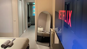 A modern hotel room with a large mirror, a bed with rolled towels, a view of a hallway, and a wall-mounted screen displaying the Netflix logo. - Maison Chic Two, a stone's throw from the Spanish Steps (Roma)