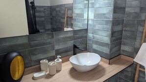 A modern bathroom with dark tiled walls, a large mirror, a white sink, and a wooden vanity. - T2 au Confort tropical downtown! (Fort-de-France)