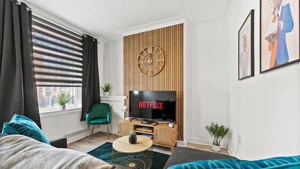 A modern living room with a large flat-screen TV, a wooden wall clock, and a cozy seating area with a green chair and a sofa. - 20% off! Boutique Doncaster Abode - Sleeps 6! (Doncaster)