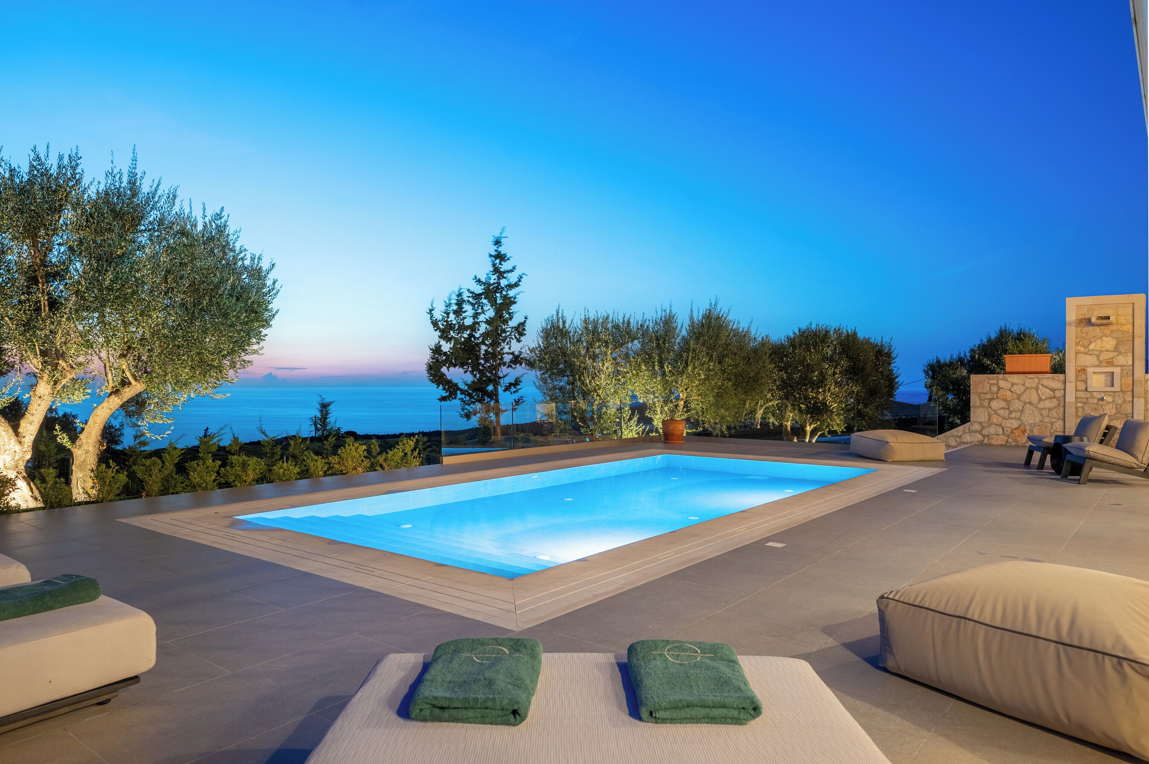 Una piscina rettangolare con cielo azzurro limpido e tramonto sullo sfondo, circondata da zone relax all'aperto e vegetazione.