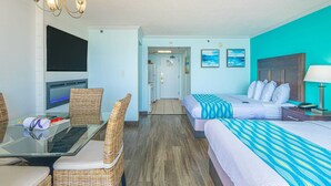 A hotel room with a TV, a dining table with chairs, two beds with striped bedding, a chandelier, and a view of the hallway. - Caravelle 712 (Myrtle Beach)