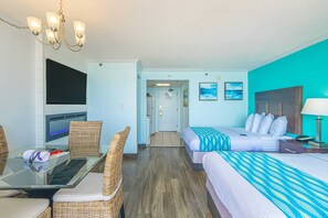 Habitación de hotel con televisión, una mesa de comedor con sillas, dos camas con ropa de cama a rayas, una araña y vistas al pasillo.