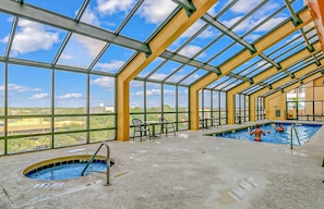 An indoor swimming pool with clear glass walls, a view of the landscape, and a few chairs and tables.