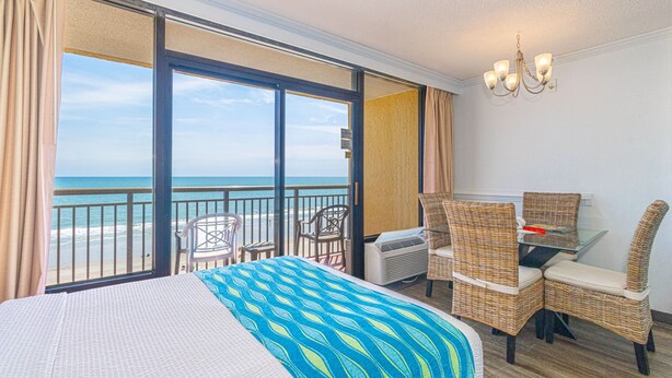 A hotel room with a large bed, a balcony with a view of the beach, wicker chairs, and a small table. - Caravelle 712 (Myrtle Beach)