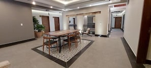 A spacious hallway with a dining area featuring a wooden table and chairs, potted plants, and a patterned rug.