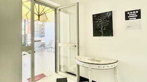 Un hall d’hôtel avec un sol à carreaux, une porte vitrée donnant sur l’extérieur, une table blanche ornée de motifs floraux et un luminaire au plafond.