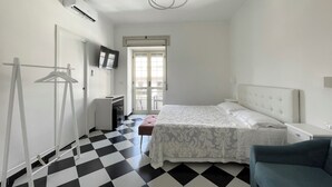 Une chambre d’hôtel avec un sol à carreaux, un lit recouvert d’une courtepointe blanche, une chaise, un bureau et un téléviseur fixé au mur.
