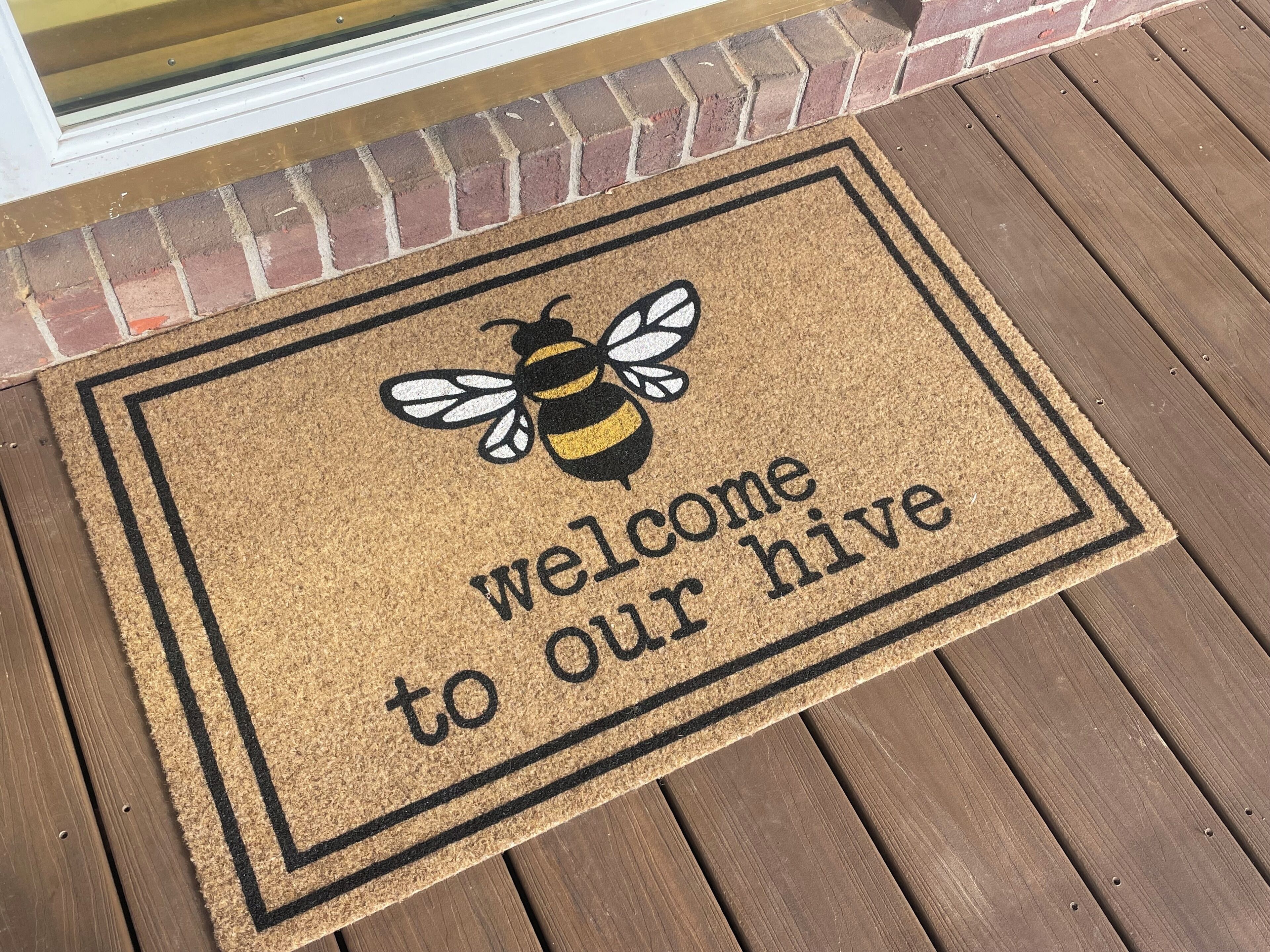 A welcome mat with a bee and the text "welcome to our hive" on a wooden deck.