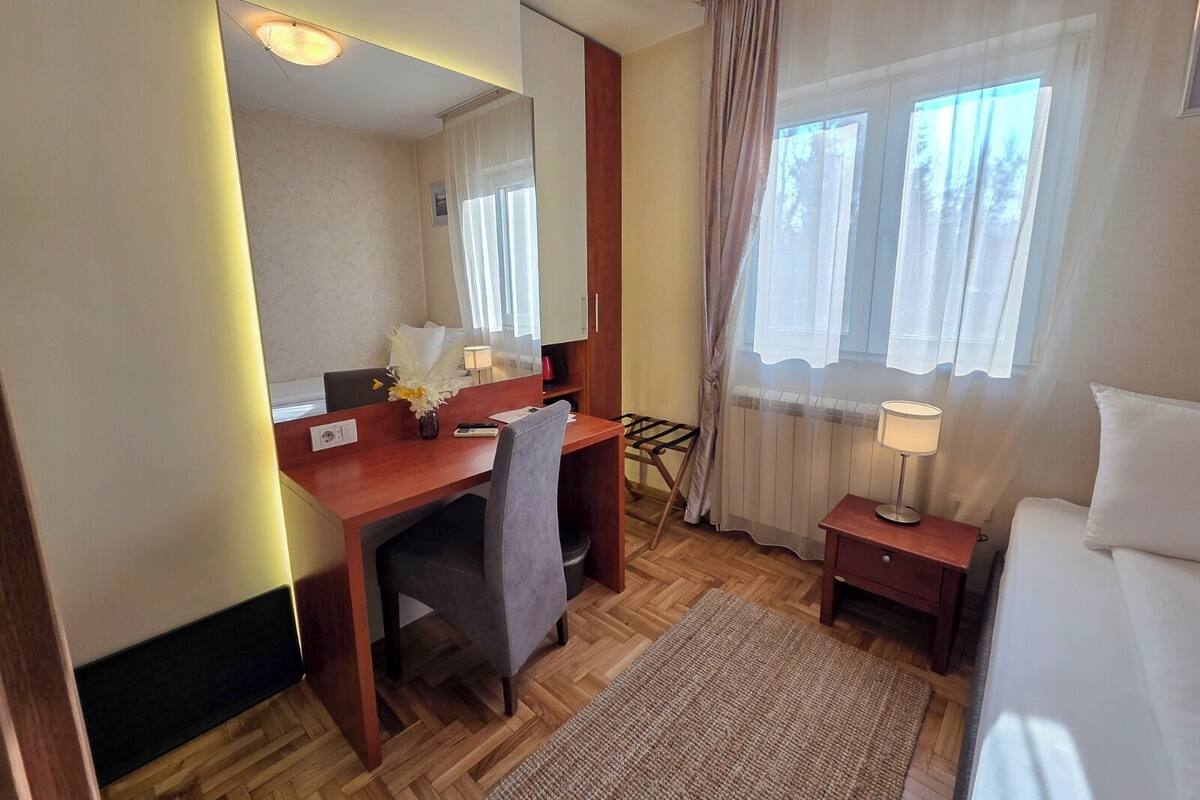 A hotel room with a wooden desk, a chair, a bed, a nightstand, a lamp, a mirror, and a window with curtains.