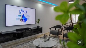 Una sala de estar moderna con un televisor de pantalla plana grande que muestra 'LOKI'S Vacation Place'. Hay una mesa de centro con un adorno, un sofá y sillones.