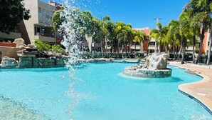 Una piscina azul cristalina con fuente, rodeada de palmeras y edificios.