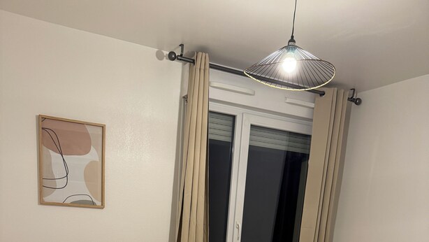 Een slaapkamer met een groot bed, een nachtkastje met twee afstandsbedieningen, een radiator en een raam met gordijnen.