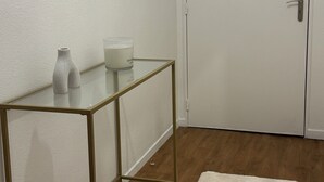 Een kamer met een witte deur, een spiegel aan de muur, een klein tafeltje met een vaas en een kaars, en een witte vloerkleed.