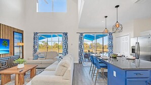 Un vaste espace de vie comprenant un canapé, une table basse et un coin repas avec des chaises bleues. On peut apercevoir une cuisine dotée d’un grand réfrigérateur et d’un îlot central bleu.