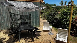 Un espace extérieur abrité avec une table et deux chaises, un bâtiment en bois en arrière-plan et un jardin avec des arbres et des arbustes.