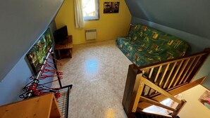 Une petite pièce mansardée avec un plafond en pente, une fenêtre avec des rideaux, un radiateur, un canapé à motifs verts, une chaise en bois et une petite table sur laquelle se trouve un téléviseur.